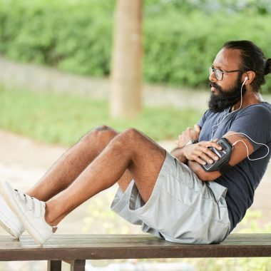 Athletic Indian man with long beard listening to music with earphones while doing abdominal crunches on bench outdoors